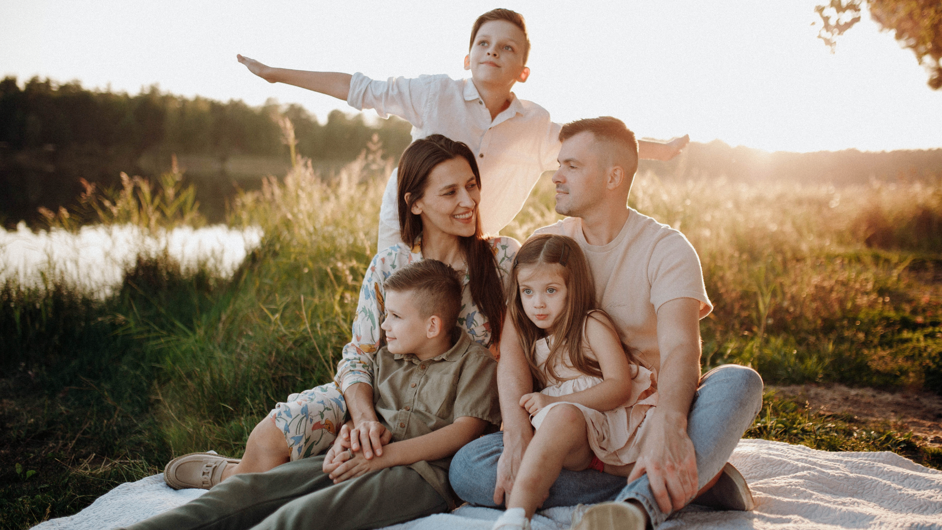 A family of five enjoying a warm sunset together by a lake — parents and three children sitting on a picnic blanket.