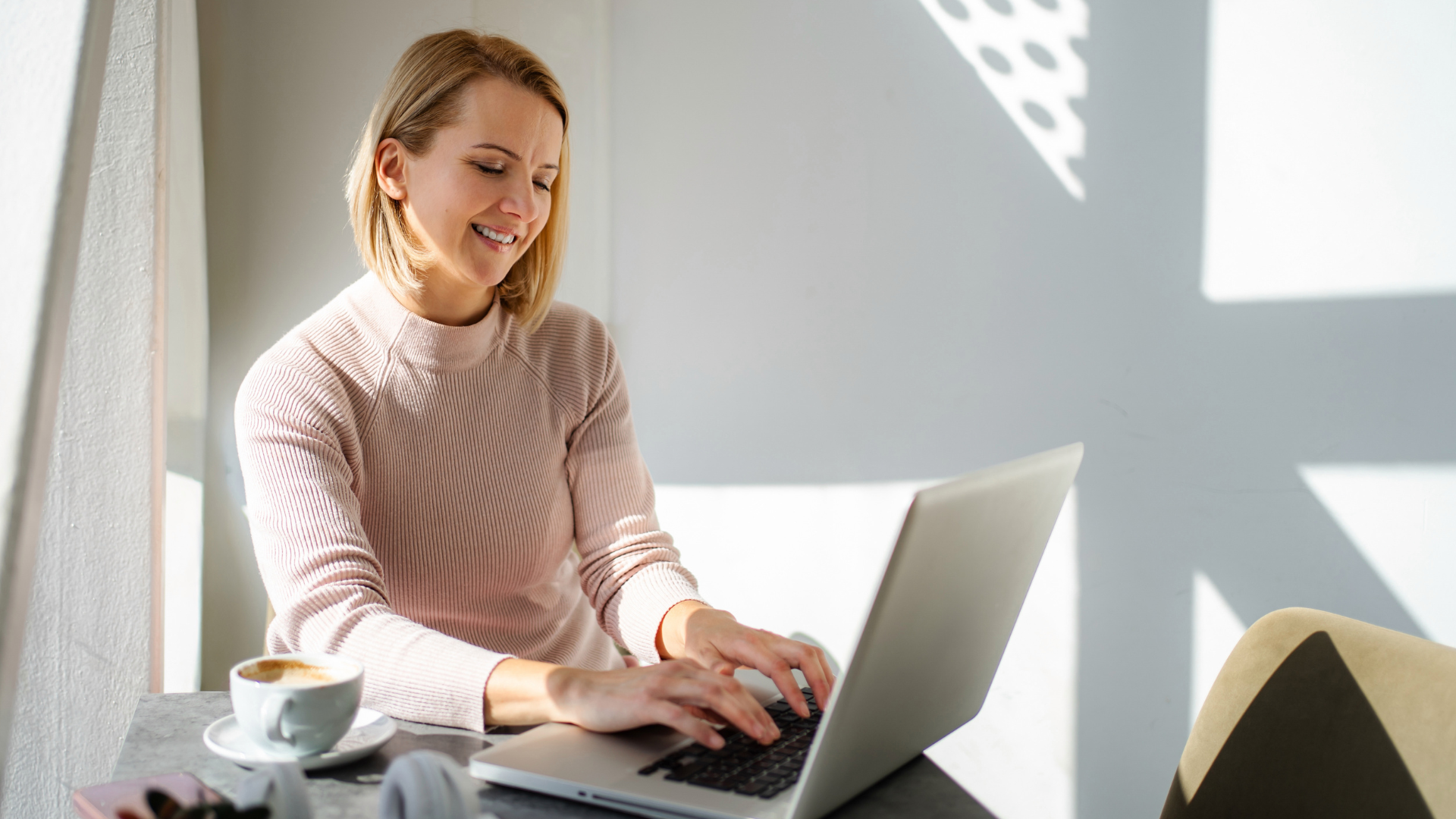 A professional smiling while working on a laptop at a sunlit table with a cup of coffee.